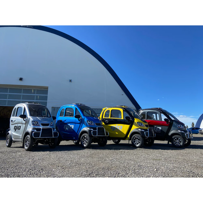 GIOEnclosedAllSeasonMobilityCar Four small electric vehicles in different colors parked in front of a building with a blue sky.