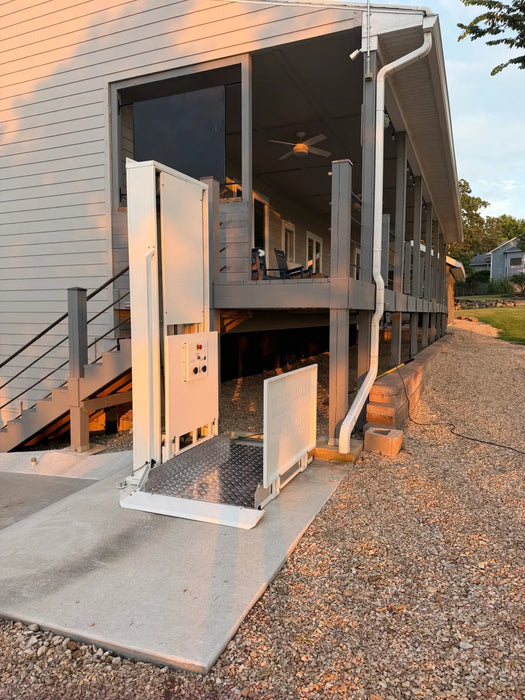 An outdoor residential wheelchai lift stairlift with an open ramp and open side door, installed at the entrance of a two-story house with a covered porch.