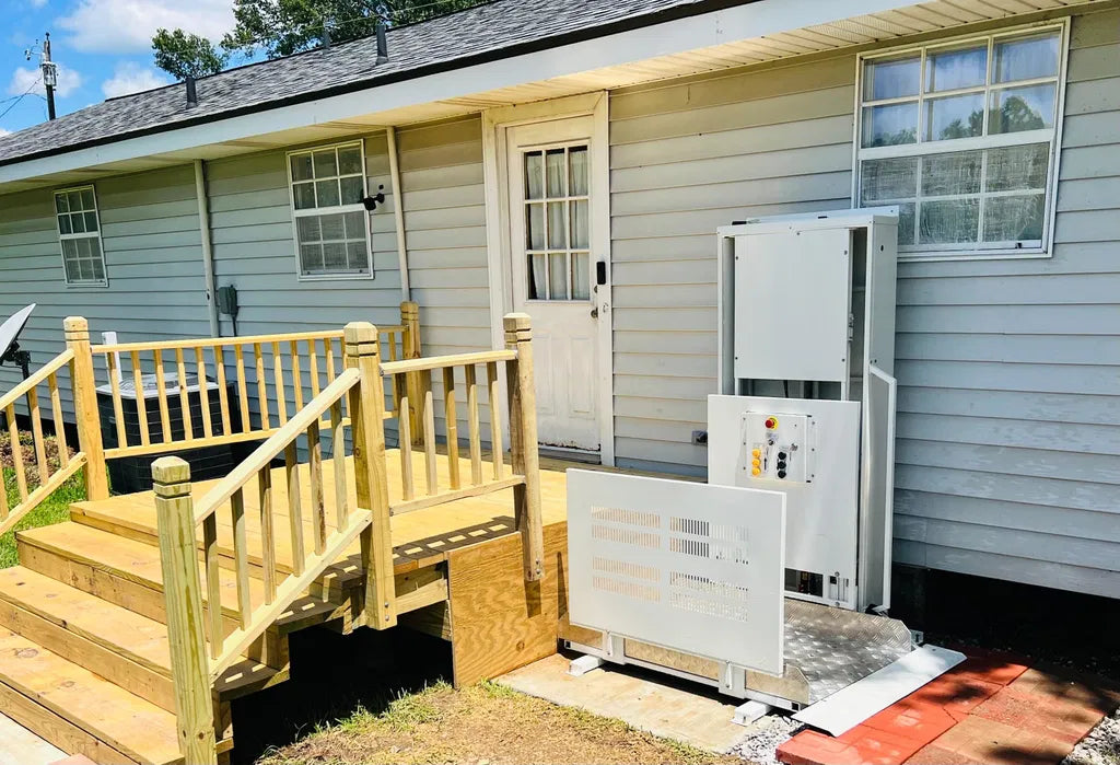 A large white outdoorPLIFT 50 residnetial wheelchair lift upositioned next to a wooden staircase and front door of a house with light gray siding.