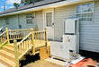 A large white outdoorPLIFT 50 residnetial wheelchair lift upositioned next to a wooden staircase and front door of a house with light gray siding.