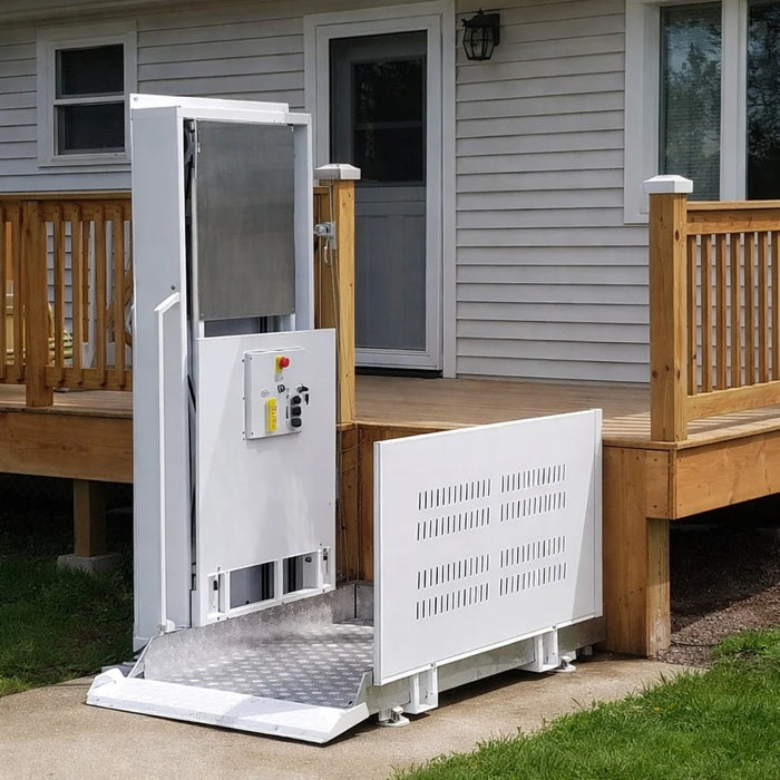 A white residential wheelchair lift with a diamond-plate metal ramp, shown installed beside a wooden deck on a house lawn.